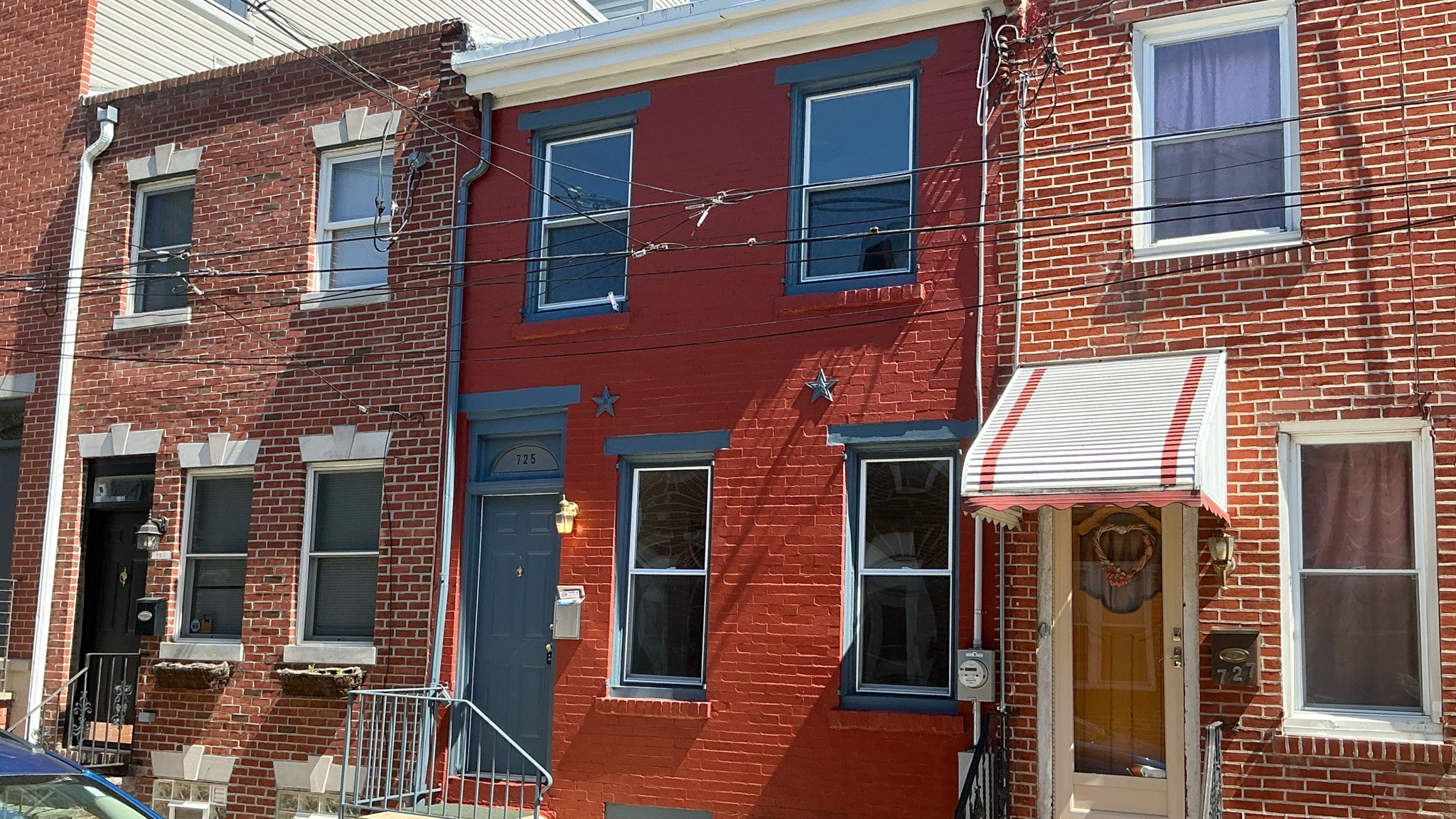 Exterior front view of a vibrant red brick rowhome in South Philadelphia, PA. The classic South Philly townhouse features bright red painted brick, contrasting blue window and door trim, a blue front door with transom, decorative star accents, metal front stoop railing, and a striped awning on the neighboring unit. Overhead utility lines and neighboring brick homes complete the iconic streetscape. Inspected April 2026