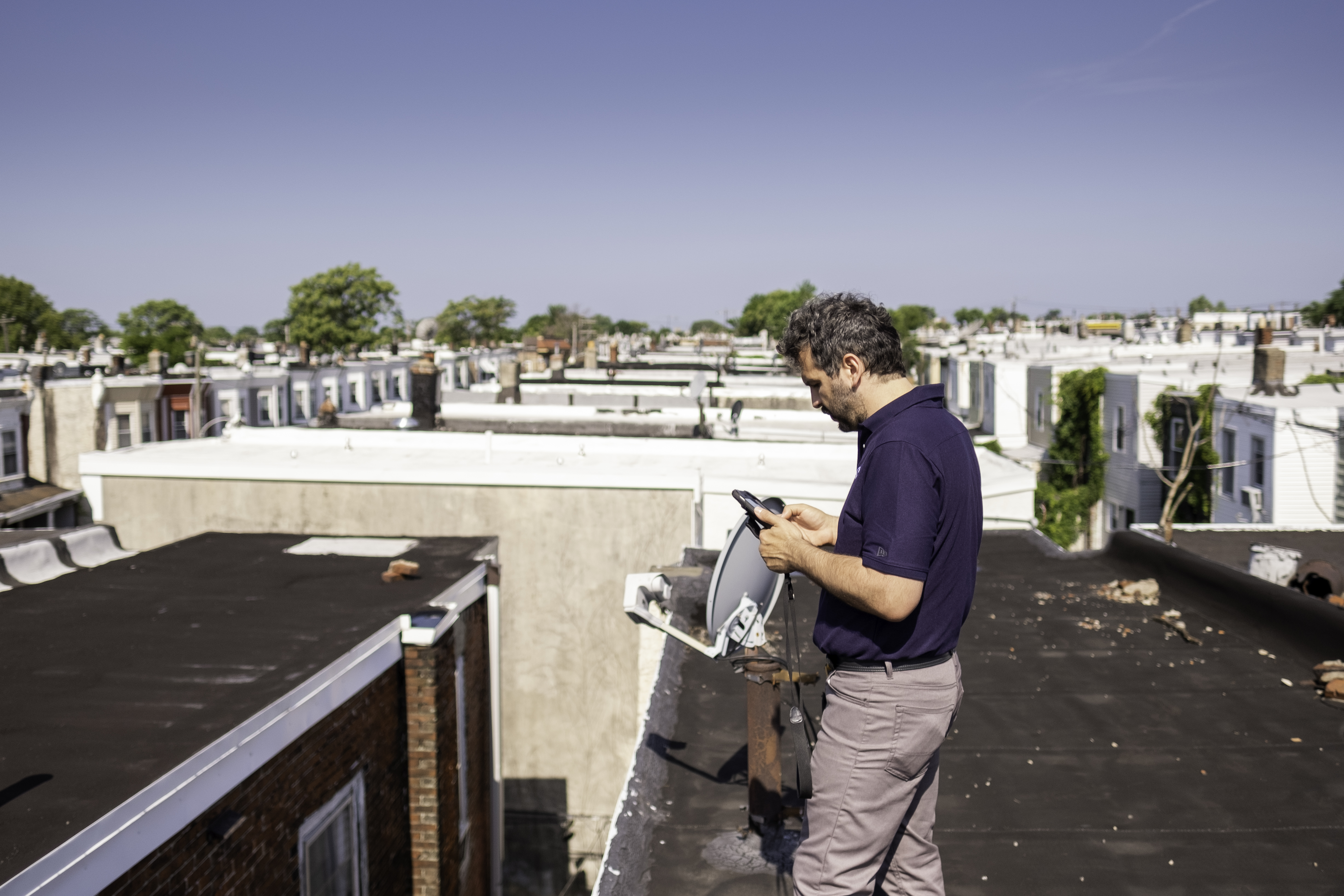 ValueGuard home inspector standing on a flat Philadelphia rowhouse roof documenting conditions on a tablet, with a block of rowhouse roofs and parapet walls behind him