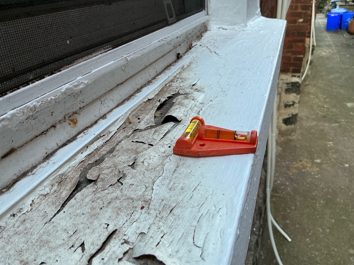 DIY home inspection photo of an exterior window sill showing severe peeling paint, cracks, and negative pitch (sloping inward toward the house) with a red level tool demonstrating the slope — high risk of moisture intrusion.