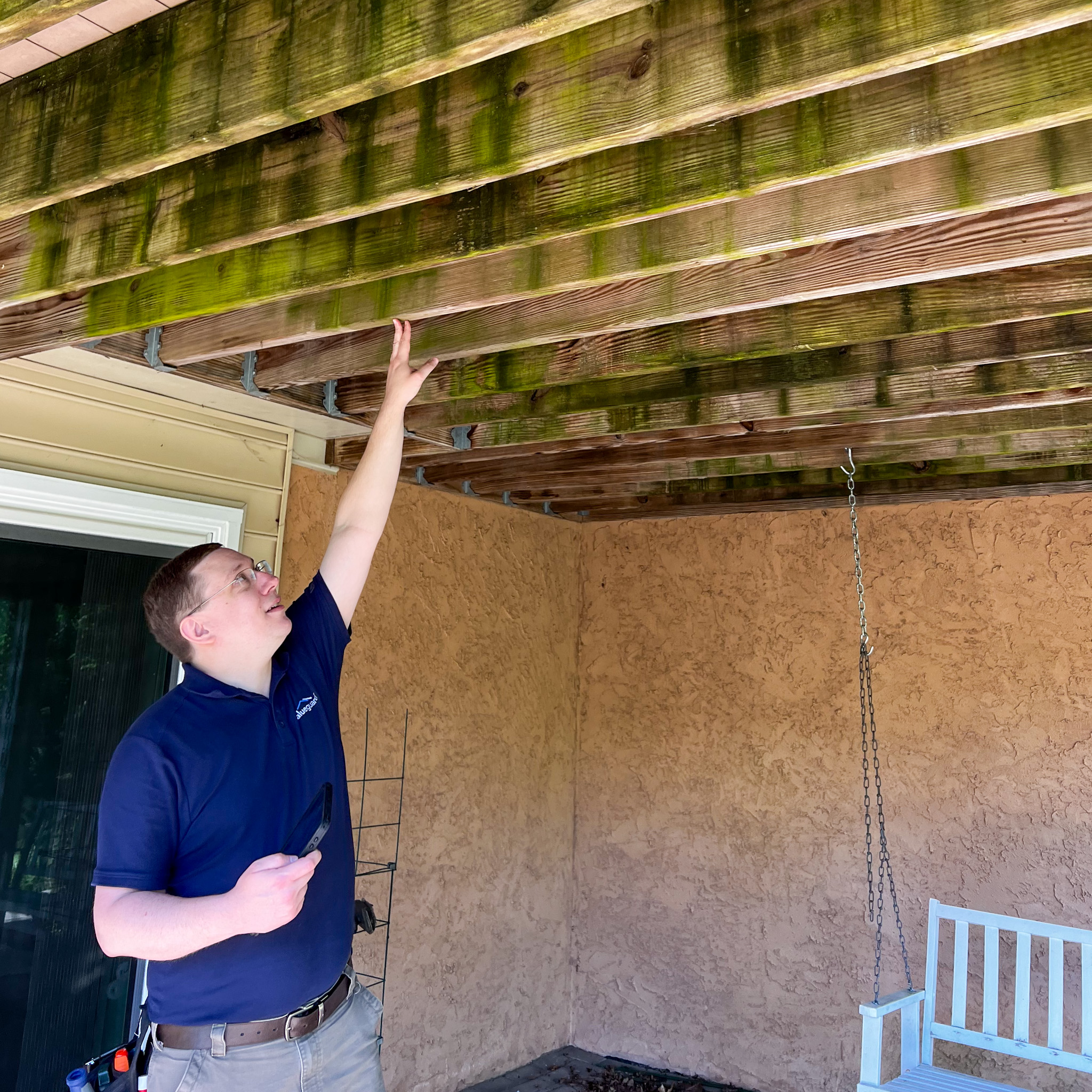 A male home inspector in a navy blue polo shirt and glasses stands under a wooden porch, reaching up with his right hand to examine covered deck joists.