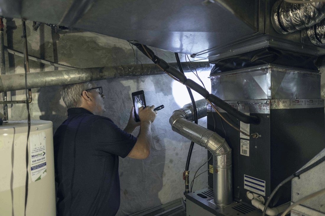 Home inspector using a flashlight and phone to examine ductwork and a furnace in a dark basement.