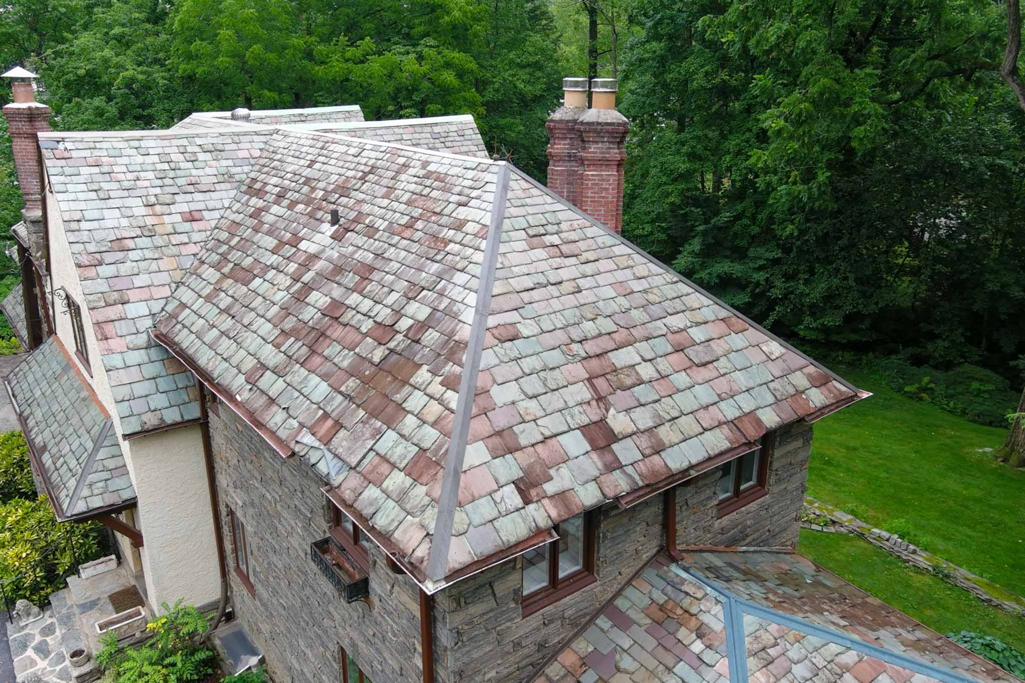 Aerial drone view of a variegated green and red slate roof with brick masonry chimneys on a 1927 stone Tudor Revival home on the Main Line