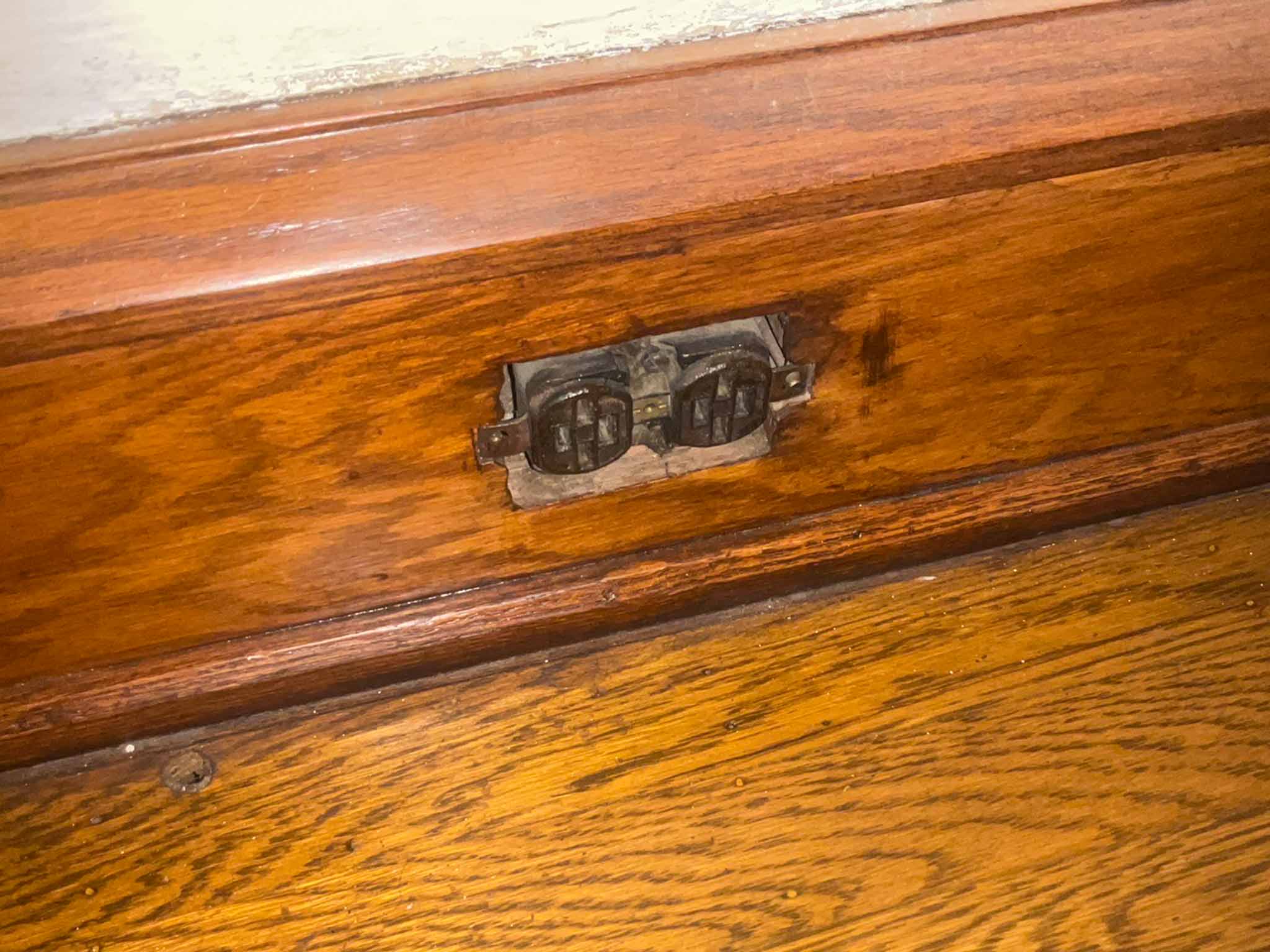 Original 1920s ungrounded two-prong electrical receptacle mounted in the oak baseboard of a Tudor Revival home found during a ValueGuard home inspection