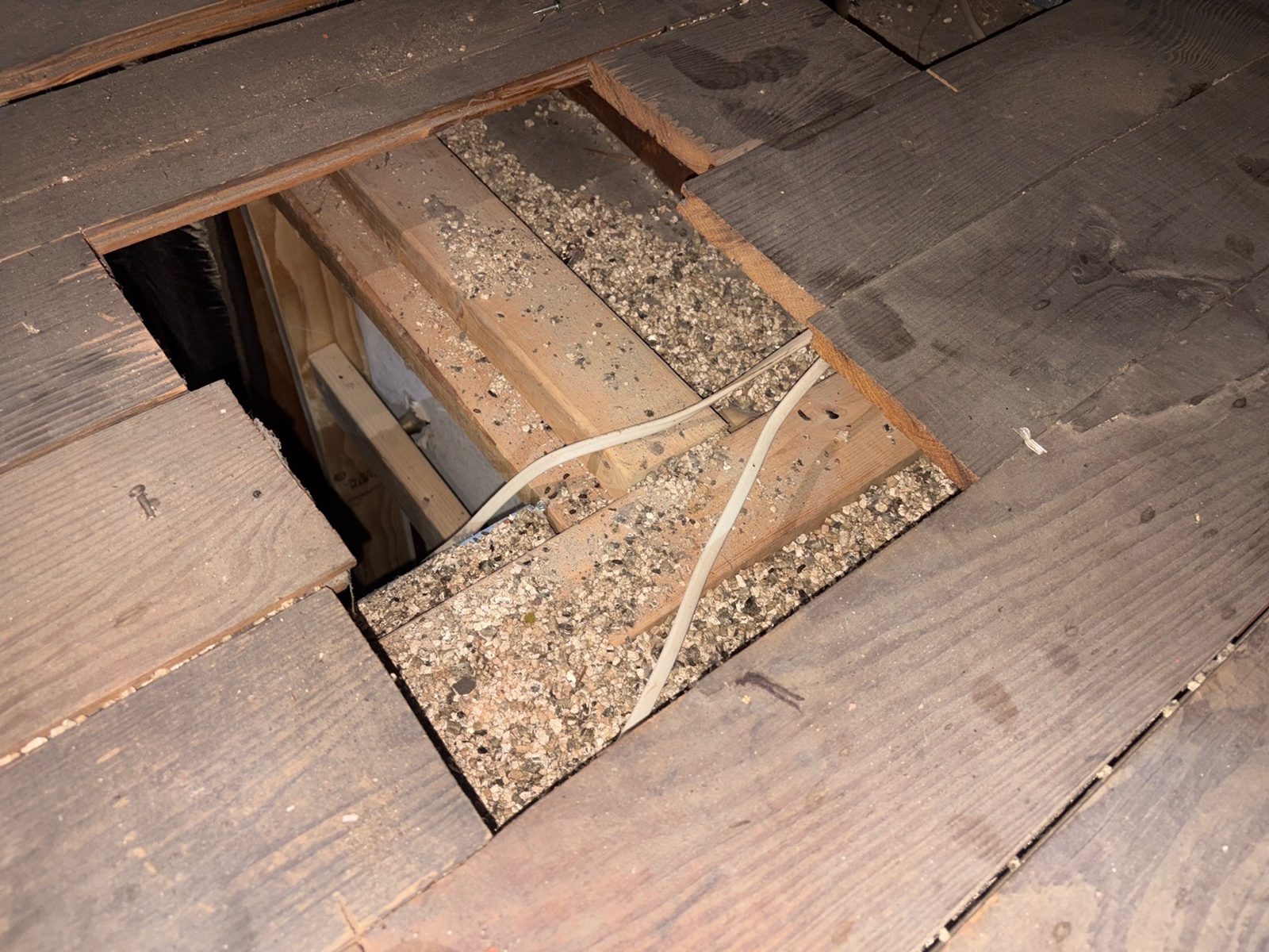 Vermiculite insulation between attic floor joists under a hole in the plank floor of a 1915 Glen Mills home, with electrical cable running through it