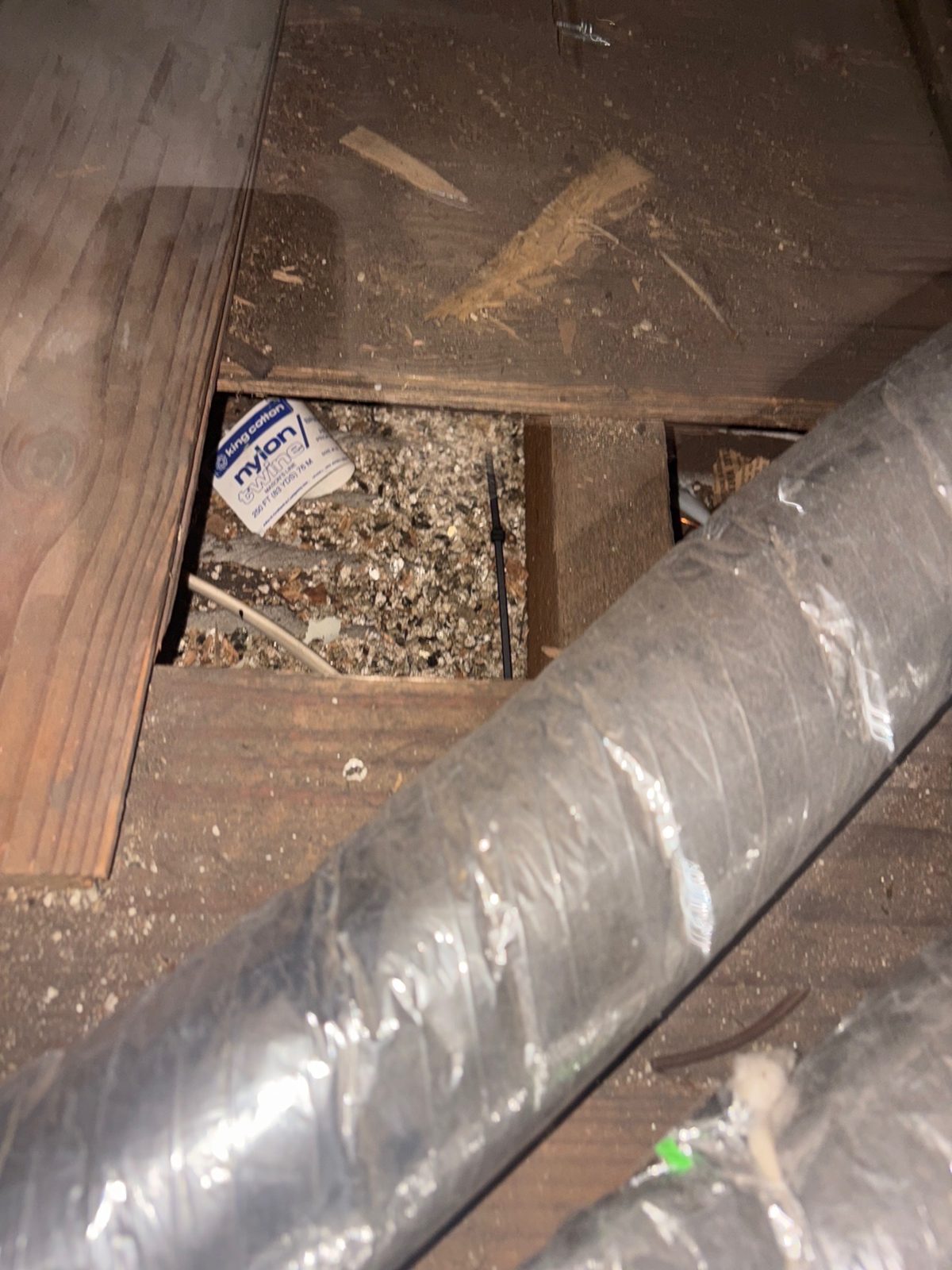 Vermiculite insulation visible between attic joists beneath a flexible HVAC duct, with old debris including a spool box of nylon twine